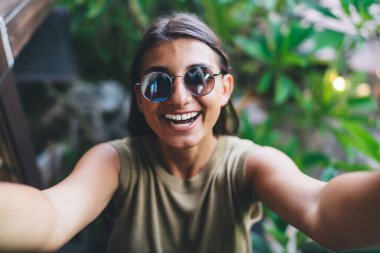 Bali tatil köyünde zaman geçirirken gülümseyen ve selfie çeken gündelik giysili ve güneş gözlüklü pozitif kadın yüksek açılı.