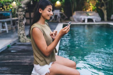 Siyah saçlı, turkuaz havuzunda akıllı telefon kullanırken gülümseyen genç ve etnik kadın turistin yan görüntüsü.