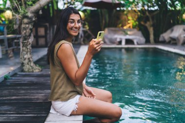 Gözlüklü iyimser bayanın cep telefonuyla mesajlaşması ve kameraya bakıp sıcak yaz tatilinin tadını çıkarması.