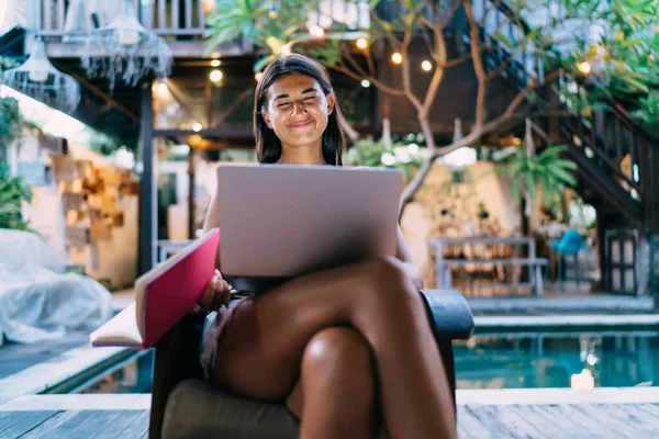 一位积极的年轻女性坐在游泳池边舒适的椅子上 一边笑一边用电脑和翻阅抄本上的笔记 — 图库照片