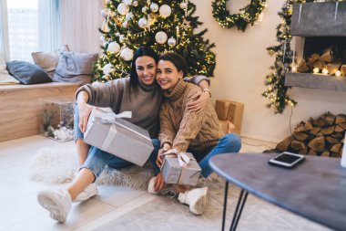 Heyecanlı kadınlar beyaz spor ayakkabılar giyip, süslü şöminenin yanındaki tahta zeminde otururken, yaylı kutularla sarılıp gülümsüyorlar.