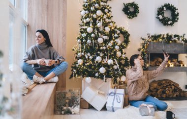 Sıcak kazaklı kadınlar kış tatili için oturma odasında dinlenirken Noel ağacına karşı selfie çekiyorlar ve çapraz bacaklı, elinde sıcak içecekle pencere eşiğinde oturuyorlar.