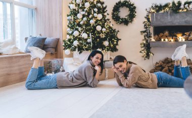 Süslü Noel ağacının yanında yerde uzanan ve kış tatilini evde birlikte geçirirken gülümseyen mutlu genç kız arkadaşlar.