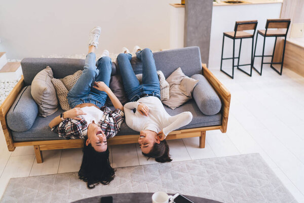 From above of full body of female friends rejoicing together while chilling on sofa in living room of modern apartment