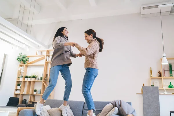 stock image Positive young female friends in warm outfits holding hands and laughing while jumping on comfortable sofa in cozy living room at home