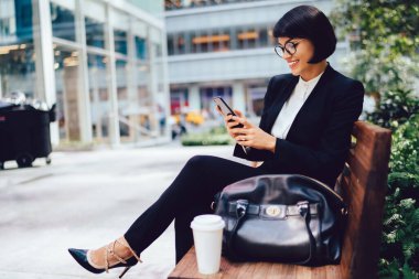 Siyah takım elbiseli ve yüksek tepeli genç bayanın parktaki ahşap bankta otururken telefonda sörf yapması.