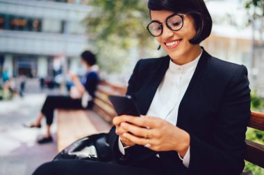 Zarif siyah takım elbiseli ve gözlüklü genç bir kadın New York 'taki bankta otururken cep telefonundan mesaj atıyor.