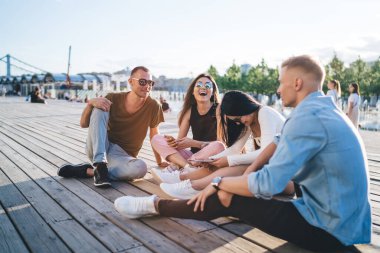 Bir grup insan, güneşli bir yaz akşamı parkta, gökyüzüne ve yeşil ağaçlara karşı ahşap zeminde otururken eğleniyor.
