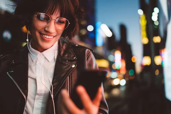Smiling hipster girl in eyewear reading message with good news from friend holding mobile smiling at night city street, pretty woman in headphones enjoying downloaded new song via application on phone