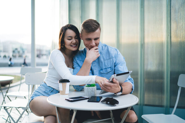 Счастливая многорасовая подруга и бойфренд в повседневной одежде, сидящие за столом и просматривающие вместе таблицу, пока пьют кофе и проводят время в кафе на открытом воздухе