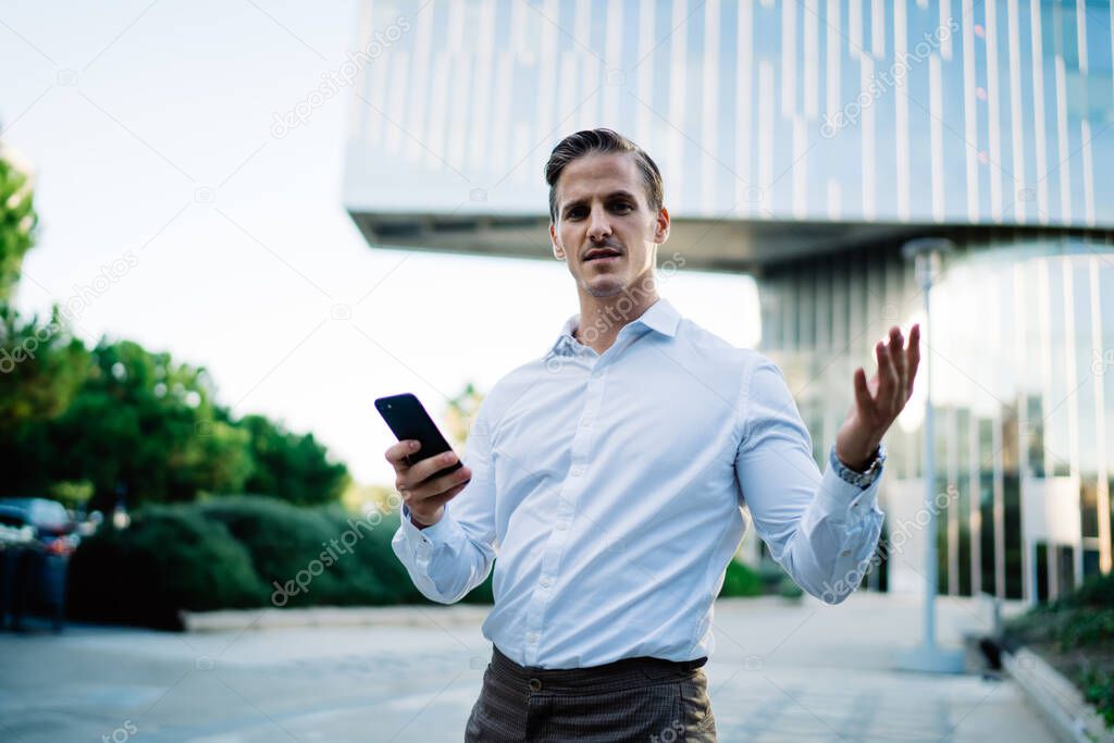 Half length portrait of puzzled male boss 30s looking at camera while waiting for international phoning via cellular, confused businessman holding modern smartphone gadget in hand and posing