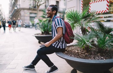 Şehirde çiçek tarhında sırt çantası ve tabletle oturan günlük giysiler içinde mutlu etnik sakallı bir erkek.