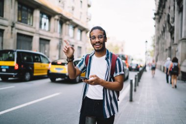 Neşeli koyu tenli hippi gezgin modern megalopolis 'te yolda duruyor. Cep telefonu taksiyi bekliyor. Şen şakrak gezgin.
