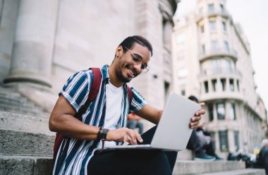 Neşeli koyu tenli Afro-Amerikalı hippi dizüstü bilgisayarda yazı yazıyor. Dizüstü bilgisayarla ve taramayla içeriği paylaşıyor. Web tarayıcıları vasıtasıyla öğrenmekten memnun olan koyu tenli olumlu erkek öğrenci.