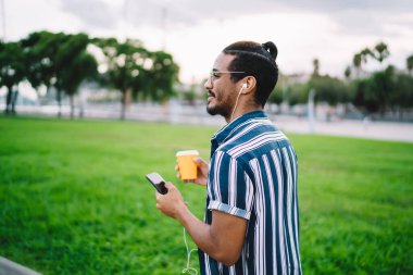 Elinde akıllı telefon, elinde kahve, elinde kahve, elinde kahve, gülümseyen Afro-Amerikan hipster adamın kulaklıkla gezip mesajlar gönderip müzik listesinin keyfini çıkardığı pozitiflerin arka görüntüsü.