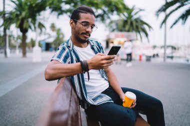 Düşünceli koyu tenli erkek etkileyicisi sokakta bankta oturan arkadaşlarıyla ve takipçileriyle iletişim kuruyor. Ciddi Afrikalı Amerikalı hipster adam cep telefonuna 4G bağlantısı üzerinden yeni bir uygulama yüklüyor.