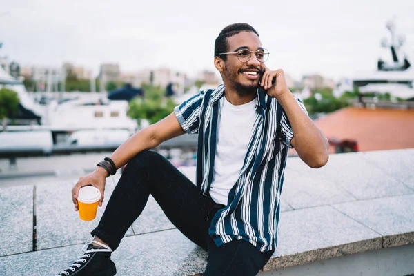一个穿着时髦服装 留着胡子的积极向上的年轻人一边坐在户外石墙边的外卖咖啡上一边用手机聊天 — 图库照片