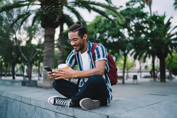Genç Afrikalı Afro-Amerikan hippi erkek rahat giyinmiş ve sırt çantalı gözlüklü akıllı telefon kullanarak taş basamakta oturuyor ve bacak bacak üstüne atmış kahvelerin keyfini çıkarıyor.