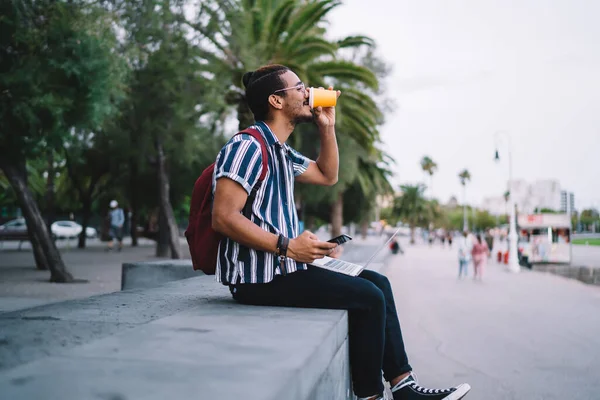 Sokakta palmiyeler arasında akıllı telefon ve dizüstü bilgisayar kullanırken kağıt bardakta kahve içen tatmin olmuş genç bir erkeğin yan görüntüsü