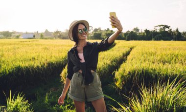 Moda güneş gözlüklü neşeli beyaz kadın selfie görüntülerine tıklamak için ön akıllı telefon kamerası kullanıyor. Vietnam 'dan internete bağlanarak seyahat videosu çeken komik bir kadın etkili.