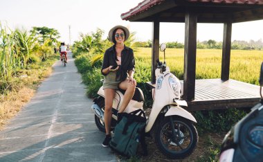 Güneş gözlüklü neşeli kadın turist portresi ve kameraya gülümseyen hasır şapkalı pirinç tarlalarının yakınındaki eski mobilet ve cep telefonuyla gülen bin yıllık hippi kız.