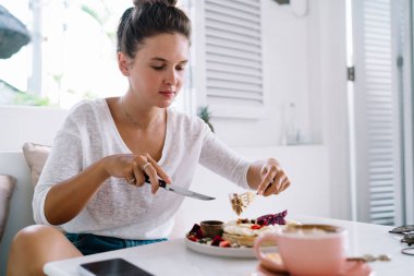 Çatal ve bıçak kullanan neşeli bir kadın öğle yemeği molasında lezzetli hamur işleri kesmek için rahat, parlak bir mutfakta tek başına oturuyor.