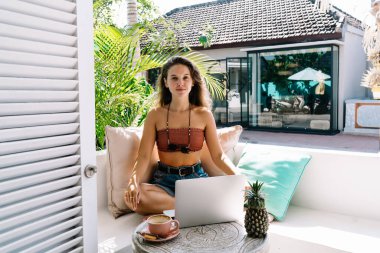 Gündelik giysiler içinde kendinden emin bir kadın bilgisayara bakarken kameraya bakıyor ve nilüfer çiçeğindeki kanepede oturuyor.