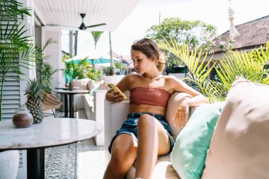 Bali 'de tatilde akıllı telefondan ilginç haberler okurken rahat bir kanepede oturan ve yastıklara yaslanan mutlu bir kadın.