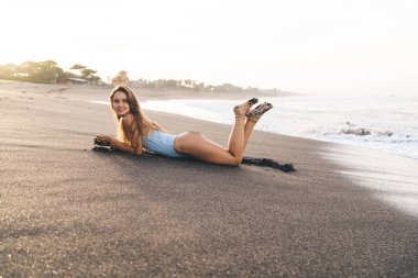 Uzun boylu, çıplak ayaklı, mayo giymiş, gülümseyen ve Bali 'de kumlu sahilde uzanmış kameraya bakan bir kadın.