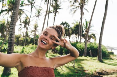 Güneşli bir yaz gününde Tayland 'da palmiye ağaçlarının yanında dururken selfie çeken ve barış jestleri yapan mutlu genç bir kadın.