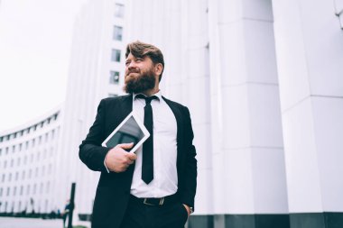 Resmi takım elbiseli yakışıklı bir girişimci. Tablet cebinde duruyor ve iş merkezinin yakınına bakıyor.