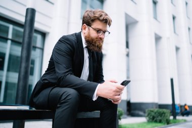 Takım elbiseli, gözlüklü ve sakallı genç bir adam bankta oturmuş internetten bilgi arıyor.