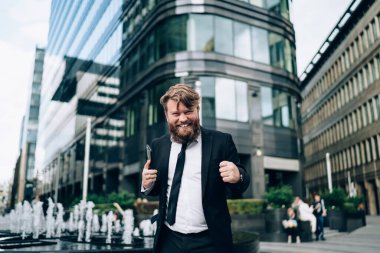 Resmi takım elbiseli ve kravatlı iyimser sakallı işadamı elinde akıllı telefonuyla modern binanın yanında duruyor ve başarılı anlaşmayı kutluyor.