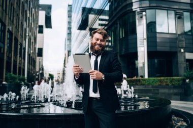 Resmi giysiler içinde neşeli sakallı bir erkek işçi. Gülümsüyor, kameraya bakıyor. Modern sokakta tablet ararken.