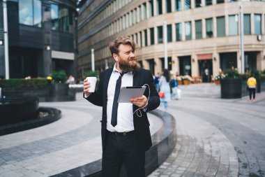 Resmi kıyafetli genç adam internet üzerinden video izlemek için tablet ve kulaklık kullanırken ofis yakınlarındaki şehir parkında dinleniyor ve içki içiyor.