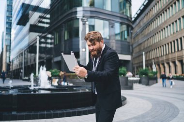 Resmi takım elbiseli, sakallı, ciddi bir genç adam şehir parkındaki tablet aracılığıyla çevrimiçi video görüşmesi sırasında konuşmaktan kafası karışmış.