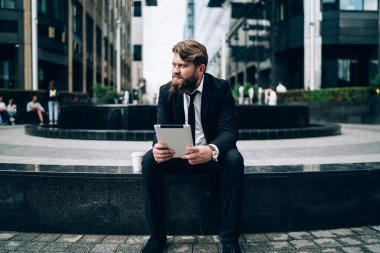 Ciddi sakallı iş adamı, resmi giysileri ve kravatıyla taş sınırında oturuyor ve tablet üzerinde çalışırken dikkatle başka tarafa bakıyor.