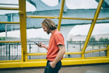 Sıradan kıyafetler içinde gülümseyen ve kulaklıklı el ele yuvarlak gözlüklü, müzik dinleyen, üstü kapalı yaya köprüsünde yürüyen akıllı telefon kullanan bir erkek.