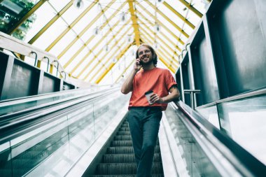 Günlük giysiler ve gözlüklerle gülümseyen, kahve içen, yaya köprüsünde yürüyen merdivenlere dayanan bir arkadaşla sohbet etmek için telefon kullanan,
