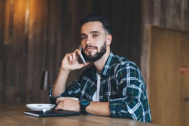 Sakallı beyaz bir adam konuşma sırasında cep telefonu ve danışmanlık cihazı kullanıyor. Kafeteryada otururken akıllı telefon uygulamasının tadını çıkaran genç bir hippi.