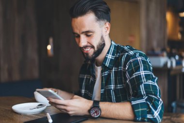 Kafkasyalı mutlu erkek blogcu kafeteryada kahve içerken komik mesajlar okuyor ve gülümsüyor, genç hippi modern akıllı telefon teknolojisiyle kamuya açık internet bağlantısı kuruyor.