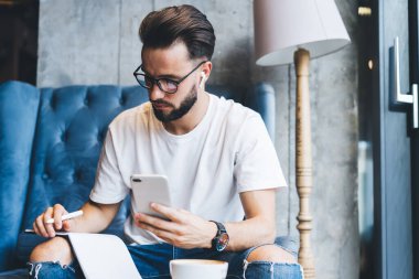 Kafkas erkek öğrenci organizasyon planı cep telefonu medya uygulamasını kullanarak boş zamanlardaki müziği dinliyor gözlüklü ve bluetooth kulaklıklı genç hippi ders kitabı notlarını kontrol ediyor.