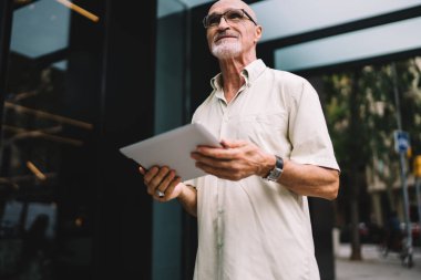 Şehir merkezindeki modern dokunmatik pedi kullanarak görüş düzeltme için klasik gözlük takan olgun Kafkasyalı yaşlı erkek blogcu 60 yaşında elinde dijital tabletle turistik caddeye bakıyor.