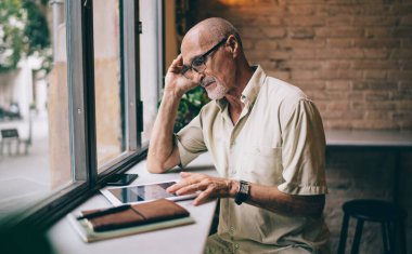 Kafe içinde boş zamanlarında sosyal medyada optik gözlük takan erkek blogger 'ın yan görüntüsü, Kafkasyalı adam 60 yaşında, çevrimiçi mesajlaşma için modern dokunmatik ped kullanıyor.