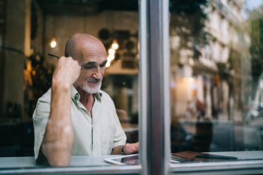 Kafkasyalı yaşlı bir adamın dijital tablet üzerinde ilginç bir ağ yayını okuduğu pencere görünümü, modern dokunmatik bloglu yaşlı bir erkek blogcu kafenin iç kısımlarında internet bilgi harcaması yapıyor