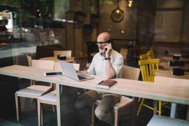 Optik gözlüklü, kafası karışmış, göz düzeltmesi için yaşlı bir adam laptopta çalışırken akıllı telefon danışmanlık konuşması yapıyor, ciddi erkek serbest çalışan 60 'lı yaşlarda video seyrediyor ve operatöre sesleniyor.