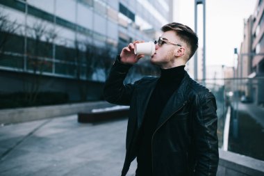 Siyah ceketli, yuvarlak gözlüklü genç bir erkek çalışanın boş bir sokakta durup içki içerken ve mola verirken yan görüntüsü.