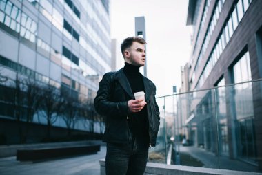 Siyah deri ceketli ve balıkçı yakalı şık genç adam şehir parkında durup kırık bir bardak içki içerken yüzünü çeviriyor.