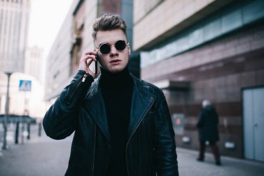 Siyah ceketli, yuvarlak gözlüklü ciddi bir genç adam caddede dikiliyor. Etrafı ofis binalarıyla çevrili ve telefonla konuşuyor.