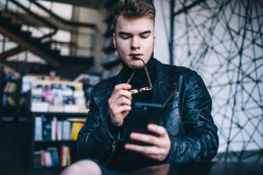 Enfes balıkçı yaka ve deri ceketli, modern kafede dinlenirken akıllı telefondan mesaj okuyan düşük odaklı genç adam.